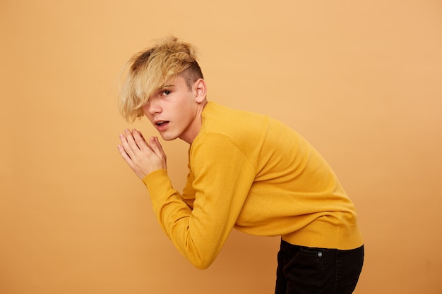 Elegante ragazzo biondo vestito con maglione giallo e jeans neri è in posa sullo sfondo beige in studio.