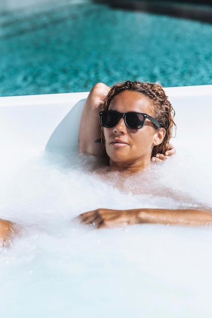 Elegante ritratto di signora che guarda sulla macchina fotografica e si diverte a fare il bagno nella vasca idromassaggio con piscina blu sullo sfondo Stile di vita delle persone di lusso Rilassamento e cura della salute del corpo Signora in vacanza estiva