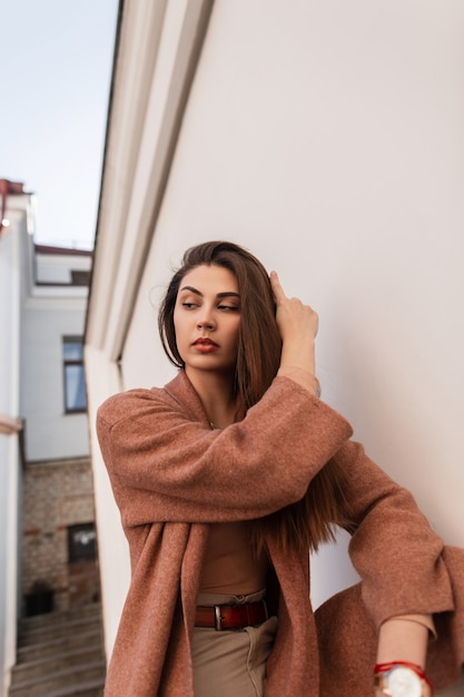 Elegante ritratto femminile bella giovane donna in cappotto alla moda con borsa in pelle marrone vicino al muro bianco vintage sulla strada. Modello di moda ragazza carina raddrizza i capelli lunghi. Signora di bellezza all'aperto.