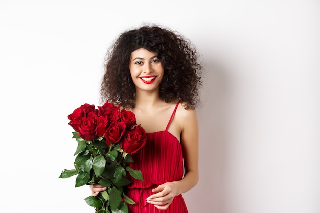 Elegante signora sorridente con labbra rosse e vestito, che tiene il mazzo di rose e che sembra felice, sfondo bianco.