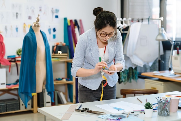 elegante stilista di moda ragazza che cattura la fotografia di abiti schizzo dal telefono cellulare in studio. sarto femminile utilizzando l'app della fotocamera del cellulare e facendo foto di disegni sul tavolo di lavoro in ufficio.