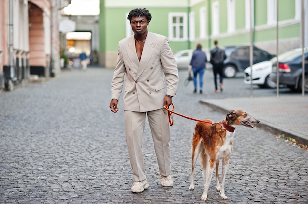 Elegante uomo afro in abito beige di vecchia scuola con cane Borzoi russo. Giovane maschio africano alla moda in giacca casual sul torso nudo.