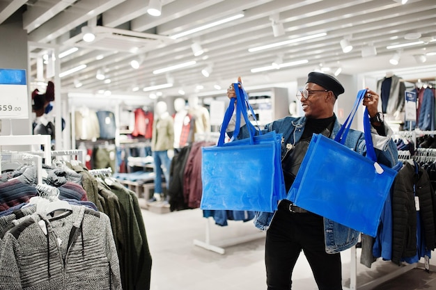 Elegante uomo afroamericano casual con giacca di jeans e berretto nero con marsupio o marsupio con borse della spesa blu al negozio di abbigliamento