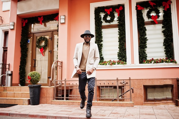 Elegante uomo afroamericano indossa giacca beige e cappello nero con occhiali da sole posa contro casa con decorazioni di Capodanno e corona.