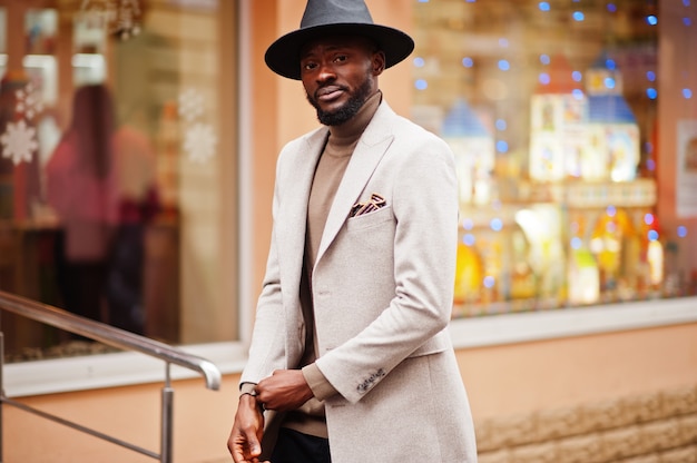 Elegante uomo afroamericano indossa giacca beige e cappello nero posa in strada.