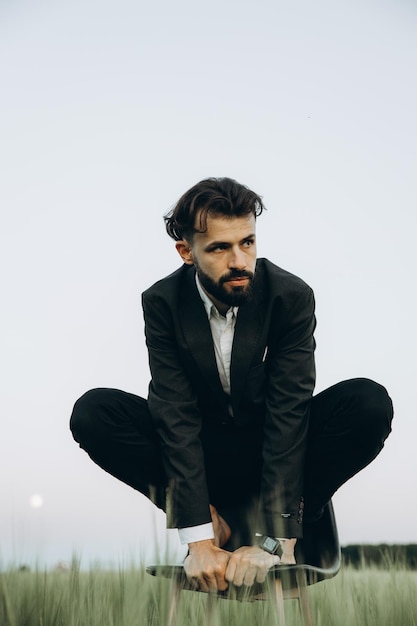 Elegante uomo barbuto in giacca nera e camicia bianca in posa per una foto nel mezzo di un campo di grano verde