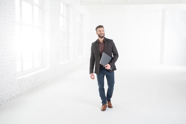 Elegante uomo d'affari in abito scuro che cammina con il computer portatile all'interno dell'ufficio bianco