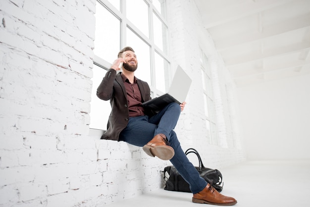 Elegante uomo d'affari seduto alla finestra con laptop e telefono cellulare all'interno dell'ufficio bianco