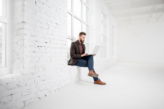 Elegante uomo d'affari seduto sulla finestra con il computer portatile nell'interno dell'ufficio bianco