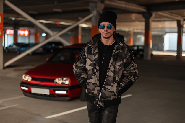 Elegante uomo hipster con occhiali da sole e cappello nero con giacca invernale militare di moda cammina per strada vicino a un'auto rossa al parcheggio