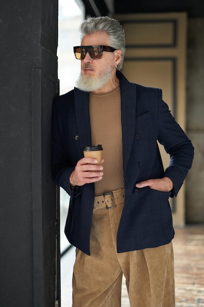 Elegante uomo maturo in abbigliamento casual da lavoro e occhiali da sole appoggiato al muro con in mano usa e getta
