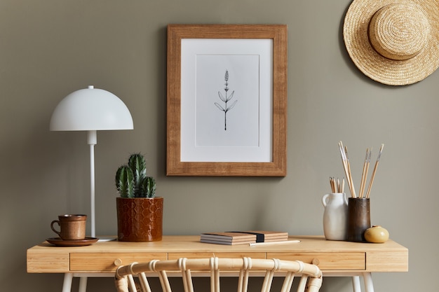 Eleganti interni bohémien di uno spazio per l'home office con scrivania in legno, poltrona in rattan, cornice per poster marrone, cactus, forniture per ufficio, cappello, decorazione ed eleganti accessori personali nell'arredamento della casa