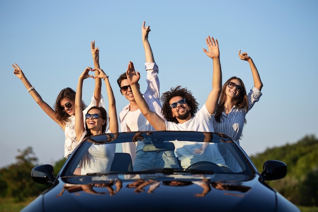 Eleganti ragazze e ragazzi in occhiali da sole sono seduti in una cabriolet nera sulla strada alzando le mani e sorridendo in una giornata di sole. .