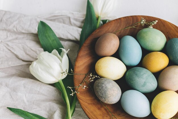 Eleganti uova di Pasqua in piatto di legno tulipani e tovagliolo di lino su tavola bianca rustica Buona Pasqua