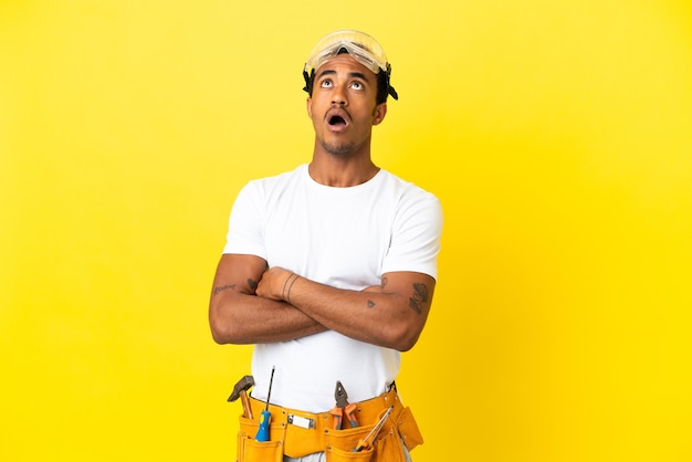 Elettricista afroamericano sopra il muro giallo isolato che guarda in alto e con espressione sorpresa