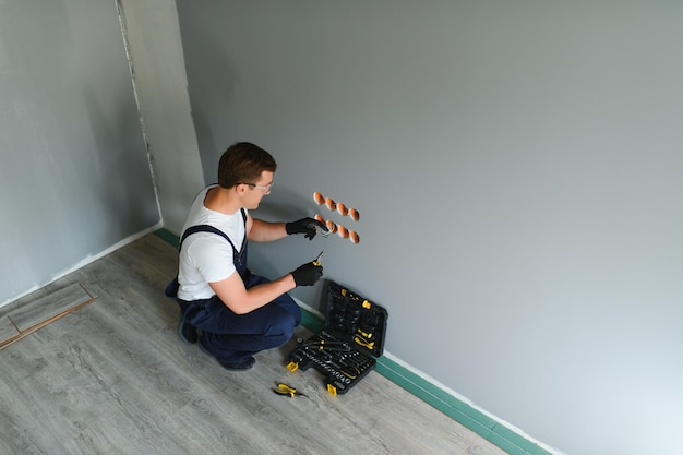 Elettricista in uniforme che monta prese elettriche sulla parete bianca all'interno