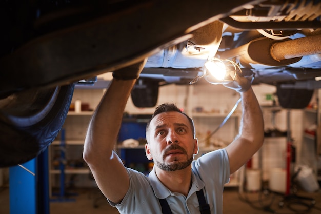 Elevato angolo di visione al meccanico maturo guardando sotto l'auto in ascensore e tenendo la luce della lampada durante l'ispezione in officina di riparazione auto, copia dello spazio