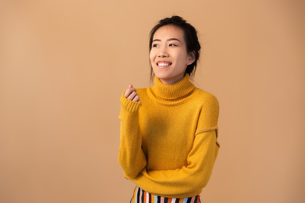 emotiva donna giapponese che indossa un maglione ridendo e guardando da parte isolato sul muro beige in studio