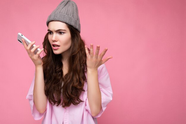 Emotivo arrabbiato attraente giovane donna bruna che indossa elegante camicia rosa e cappello grigio isolato su sfondo rosa tenendo in mano e utilizzando il telefono cellulare comunicando e registrando un messaggio vocale.