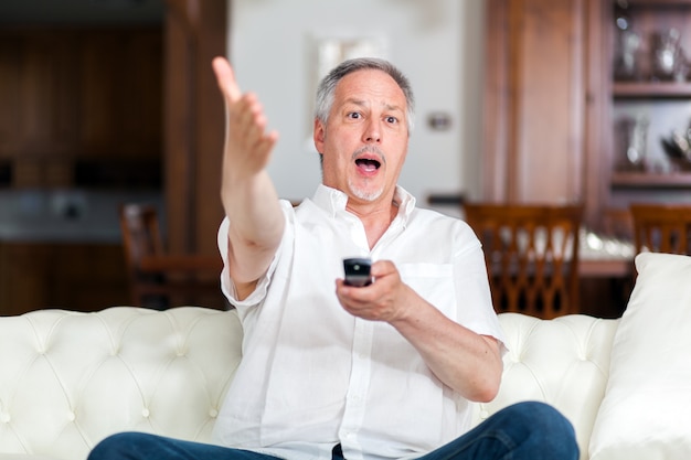 Emotivo uomo anziano guardando la tv, seduto sul divano con telecomando, copia spazio