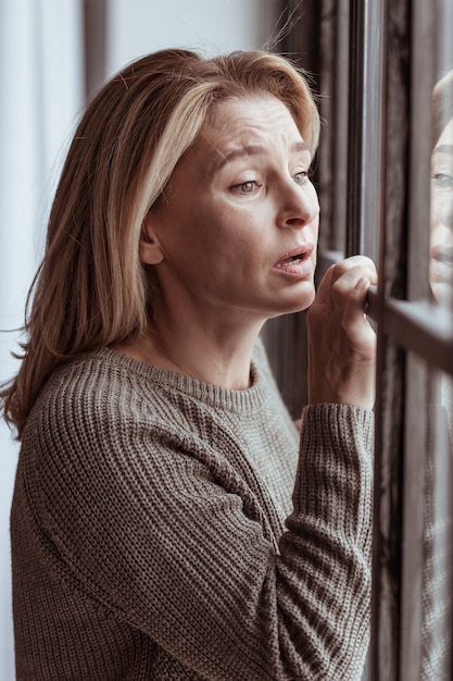 Emozionante e triste. Donna matura dagli occhi verdi che si sente emotiva e triste in piedi vicino alla finestra