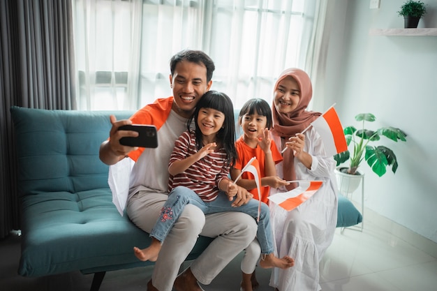 Emozionata famiglia asiatica musulmana che fa selfie e videochiamata usando il telefono a casa mentre tiene in mano la bandiera indonesiana