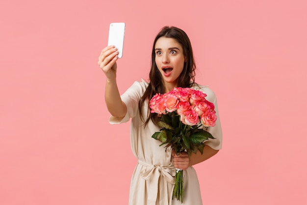 Emozioni, bellezza e concetto di romanticismo. Attraente donna bruna eccitata e seducente con belle rose, fiori ricevuti e selfie con espressione sorpresa stupita, rosa