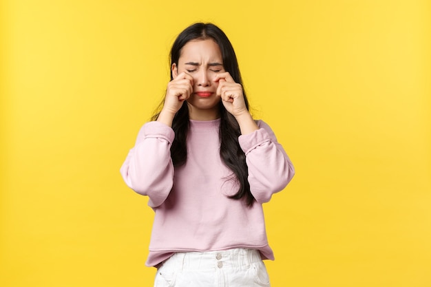 Emozioni della gente, stile di vita e concetto di moda. Ragazza asiatica triste e turbata che singhiozza, piange e si asciuga le lacrime dal viso, sentendosi depressa, in piedi sullo sfondo giallo.