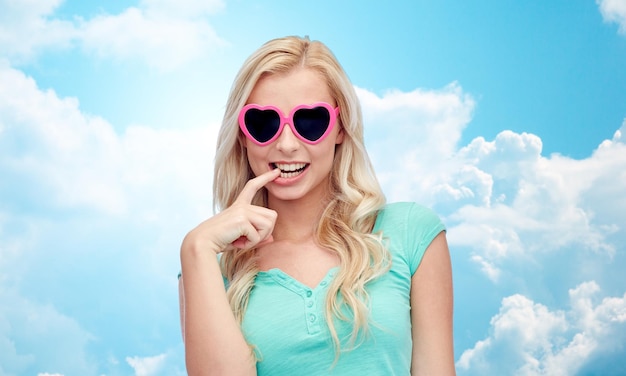 emozioni, espressioni, estate e concetto di persone - sorridente giovane donna o ragazza adolescente in occhiali da sole a forma di cuore su sfondo blu cielo e nuvole