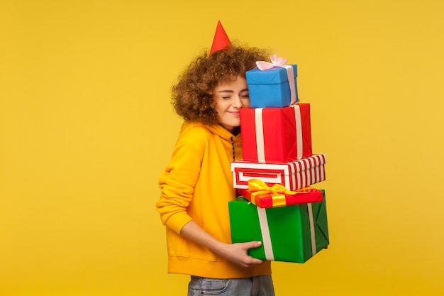 Emozioni felici dalla grande montagna di regali Ritratto di donna dai capelli ricci felice felice con cono di festa che abbraccia molte scatole presenti che celebrano il compleanno studio girato isolato su sfondo giallo