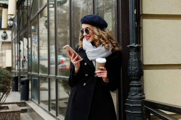 Emozioni, persone, bellezza e concetto di stile di vita - Ragazza francese. Foto di strada giovane donna che indossa abiti classici alla moda. Concetto di moda femminile. stile francese. Ragazza che manda un sms allo smartphone. caffè