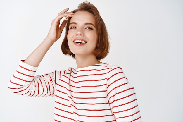 Emozioni positive e felicità. Bella giovane donna che sorride e ride, tocca il taglio di capelli con la punta delle dita, in piedi felice contro il muro bianco