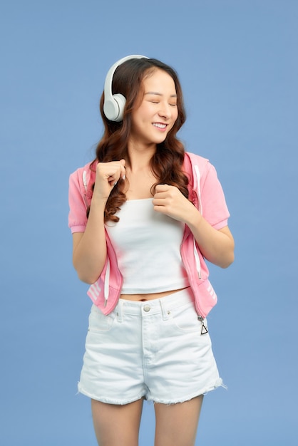 Energia ragazza asiatica con le cuffie che ascolta la musica con gli occhi chiusi su sfondo blu in studio. Indossa una maglietta bianca, pantaloncini.