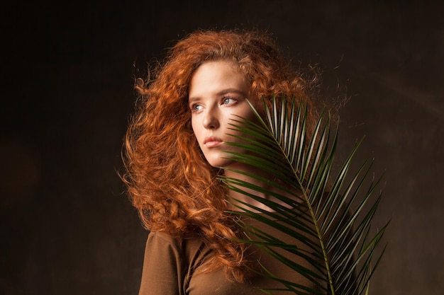 Energizer bella giovane donna con capelli ricci allo zenzero con ramo di palma verde pianta tropicale posto in studio di pareti scure Copia spazio sullo sfondo