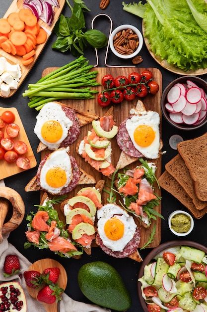 Enorme colazione sana con panini con uova strapazzate, salsiccia, salmone, rucola, ricotta, avocado su una tavola di legno, pomodori secchi