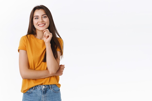 Entusiasta bella ragazza fortunata di successo in maglietta gialla tenere il mento in alto e sorridere sicuro di sé toccando la mascella sentendosi fortunato e determinato a vincere in piedi soddisfatto sfondo bianco