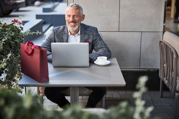 Entusiasta uomo invecchiato in un blazer elegante che sorride mentre è seduto a un tavolo da pranzo con il suo laptop e caffè