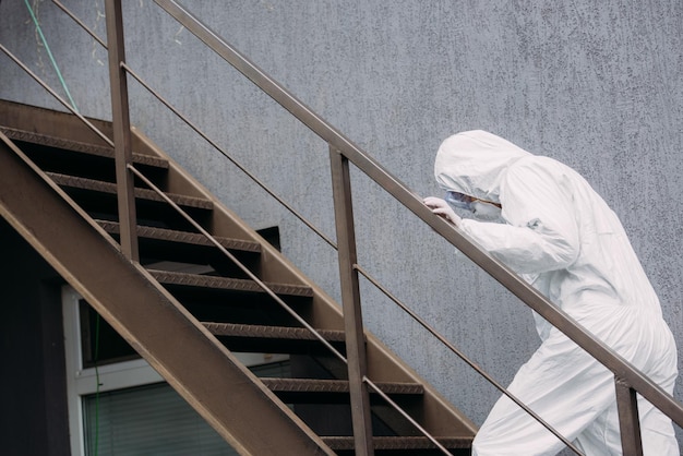 Epidemiologo asiatico in tuta ignifuga e maschera respiratoria che cammina al piano di sopra fuori dall'edificio