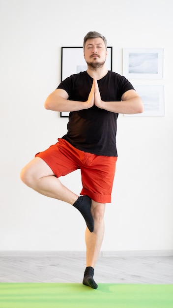 Equilibrio yoga Uomo calmo Pratica domestica Meditazione armonia Ragazzo sportivo ispirato in abbigliamento sportivo casual che rimane posa albero tenendo le mani namaste nell'interno della stanza luminosa
