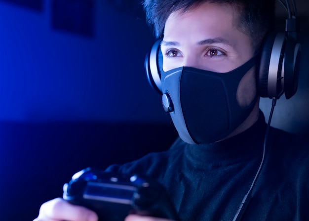 Equipaggi il gioco del video gioco che indossa una maschera chirurgica, con le cuffie. Concetto: autoisolamento per coronavirus (COVID-19) è tempo di giocare ai videogiochi.