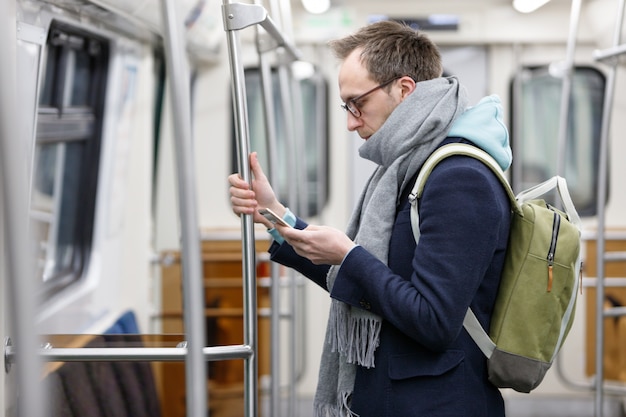 Equipaggi il passeggero in occhiali con il corrimano della tenuta dello zaino, usando ed esaminando il telefono cellulare astuto al treno sotterraneo