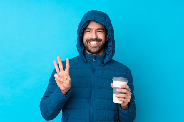 Equipaggi il rivestimento invernale da portare e la tenuta del caffè asportabile sopra la parete blu isolata felice e contando tre con le dita