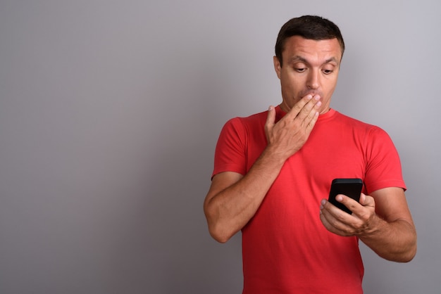 Equipaggi la camicia rossa d'uso mentre usando il telefono cellulare contro la parete grigia