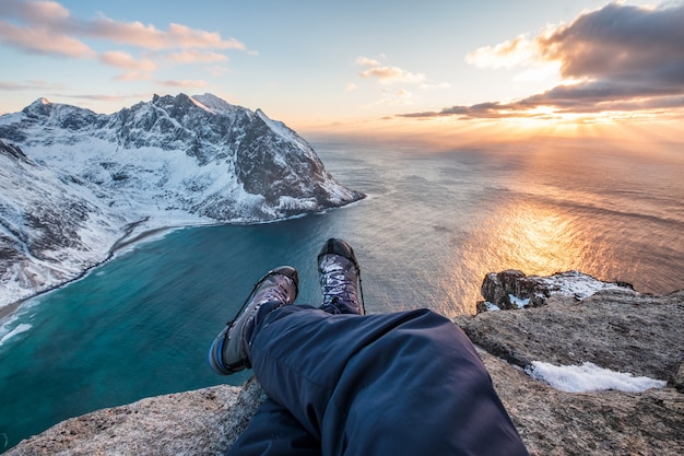 Equipaggi le gambe trasversali della viandante che si siedono sulla montagna di punta con la linea costiera al tramonto