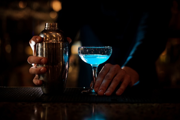 Equipaggi le mani che tengono un vetro con il cocktail e la bottiglia blu dell'estate