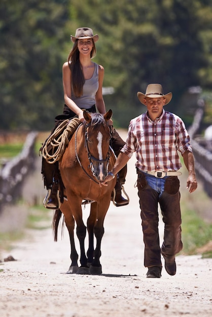 Equitazione 101 Un'attraente giovane cowgirl a cavallo in un ranch mentre un cowboy cammina a fianco