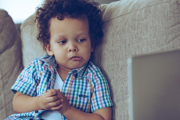 Era inaspettato! Piccolo bambino africano che guarda il suo laptop e sembra confuso mentre è seduto sul divano di casa