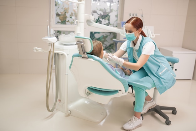 Esaminando la ragazza. Dentista bambino dai capelli rossi che indossa l'uniforme che esamina la ragazza al mattino