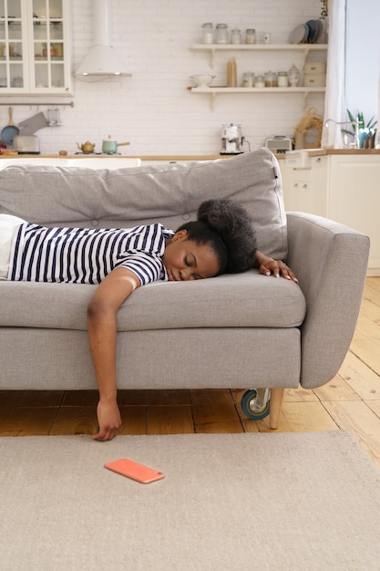 Esaurita donna afro-americana che dorme sul divano a casa, lasciando cadere il cellulare sul pavimento. Fatica