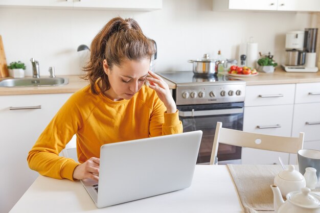 Esaurito stanco donna che lavora utilizzando il laptop tenendo la testa in mano seduto in cucina a casa